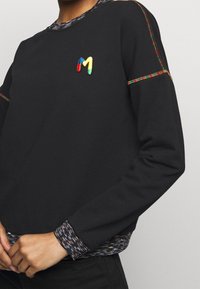 Camisa negra de manga larga con un logo "M" multicolor bordado en el pecho, costuras de arcoíris y puños en capas con patrones.