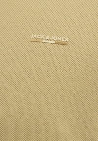 Beige textil, amely textúrált mintával rendelkezik, és arany "JACK & JONES" logó címke van belevarrva a szövetbe, középen, a teteje közelében.