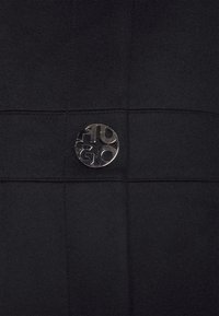 Sort stof med vandret og lodret syning har et cirkulært metalemblem med teksten "HUGO" præget.