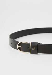 Ceinture en cuir noir avec une boucle en métal doré, dotée d'un passant marqué et de plusieurs trous de taille. Texture lisse et design classique.
