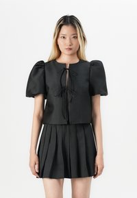 Svart cropped topp med pufferm og frontknute, kombinert med en plissert svart skjørt, alt i glatt stoff. Minimalistisk design.