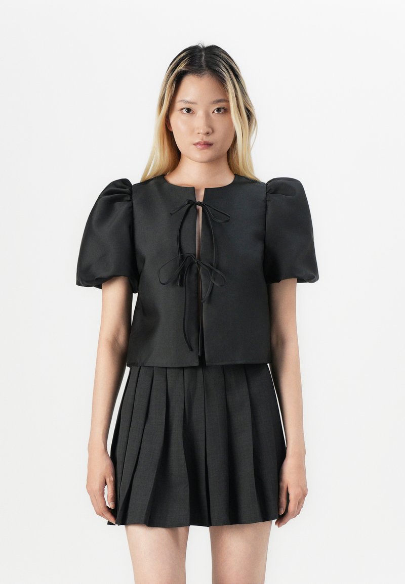 Svart cropped topp med pufferm og frontknute, kombinert med en plissert svart skjørt, alt i glatt stoff. Minimalistisk design.