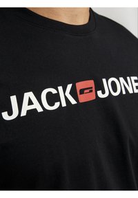 Camiseta negra con el texto blanco "JACK JONES" y un logo cuadrado rojo impresos en el pecho, llevada por una persona.