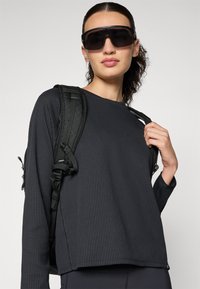 Black ribbed long-sleeve top with a crew neckline, paired with a textured black backpack and large black sunglasses.