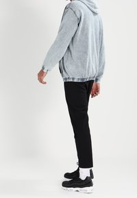 Hellblaue Denimjacke mit Kapuze, elastischen Bündchen und Saum; kombiniert mit schwarzen schmal geschnittenen Hosen und schwarzen Turnschuhen mit weißen Akzenten.