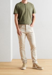 Camisa henley de manga corta en verde oliva, jeans beige y zapatillas beige. Diseño de corte recto con detalles de botones y logo en contraste en las zapatillas.