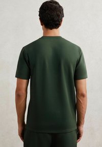 Tricou scurt cu mâneci, de culoare verde închis, realizat dintr-un material neted, având un gât clasic și o croială normală, văzut din spate.