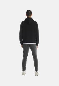 Musta hupullinen collegepaita väljällä istuvuudella, yhdistetty harmaisiin kapeisiin juoksuhousuihin ja valkoisiin lenkkareihin, nähtynä takaa.