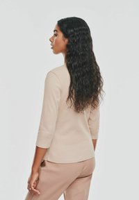 Beżowa bluzka z rękawem 3/4 z prążkowanej tkaniny, zestawiona z jasnobezowymi spodniami. Długie, falujące ciemne włosy spływają na plecy. Prosty, gładki design.