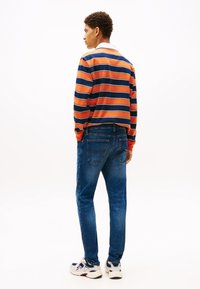 Mees, kes kannab oranži ja tumesinise triibulise pikavarrukaga särki, siniseid teksaseid ja valgeid tossusid, seisab seljaga vaataja poole tavalise tausta ees.