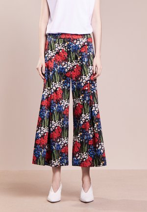Pantalones de pierna ancha con estampado floral en rojo, azul y blanco sobre tela negra, combinados con una blusa blanca y zapatos blancos de punta.