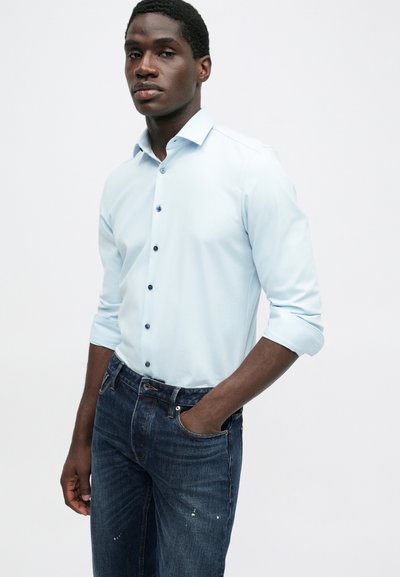 Mann iført lys blå skjorte med knapper og oppsydde ermer, og mørkeblå jeans, stående med den ene hånden i lomma.