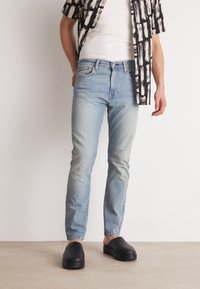 Ljusblå denimjeans med en tapered passform, kombinerat med en vit linne och en mönstrad svart-vit skjorta, samt svarta slip-on skor.