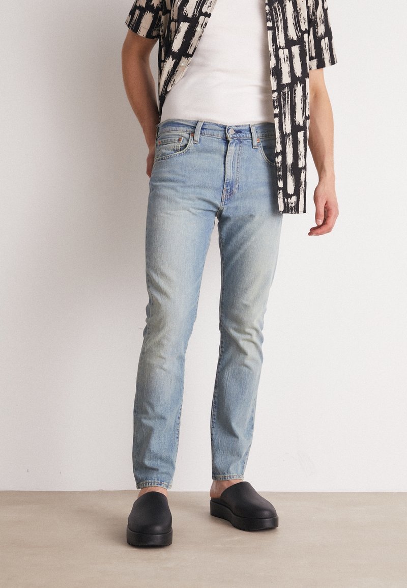 Ljusblå denimjeans med en tapered passform, kombinerat med en vit linne och en mönstrad svart-vit skjorta, samt svarta slip-on skor.