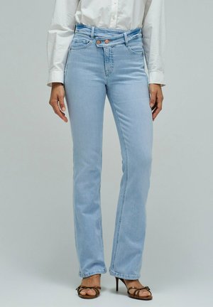 Femme portant un jean droit taille haute bleu clair, une chemise blanche à manches longues et des sandales à talons hauts marron, debout sur un fond gris.