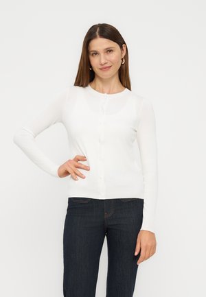 Frau mit glattem braunem Haar, die goldene Creolen, ein weißes, langärmliges, geknöpftes Oberteil und dunkelblaue Jeans trägt, mit der Hand in der Hüfte.