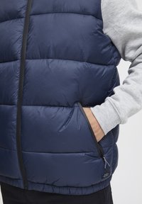 Sotilasnavy sininen pufferehka, jossa on vaakasuorat tikkauslinjat, musta vetoketju ja sivutasku. Käytetään harmaan pitkähihainen paidan päällä. Sujuva, eristävä rakenne.