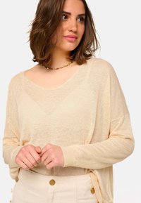 Pull oversized beige clair en tricot transparent et texturé, avec un large col en V et des manches longues. Associé à un pantalon beige taille haute.