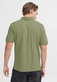 Polo vert olive à manches courtes, col côtelé et petit détail d'étiquette près de l'ourlet. Tissu simple et texturé.