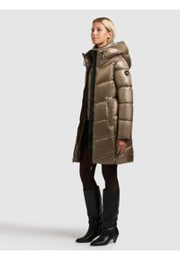 Vadderad lång kappa i metallisk olivgrön färg, med huva, dragkedja och sidofickor. Bärs med svarta knähöga stövlar.