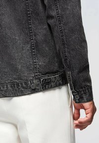 Giubbotto in denim nero con texture leggermente sbiadita, caratterizzato da dettagli con bottoni sui polsini e un design liscio e strutturato.