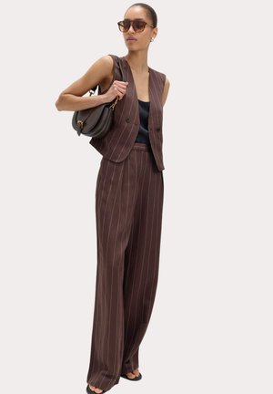 Kvinde iført brun pinstripe vest og matchende vidde bukser med sort top, solbriller og bærende en mørkebrun håndtaske over skulderen.