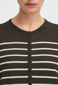 Mørkebrun cardigan med horisontale hvite striper, ribbet hals, og frontknapper i matchende brun nyanse. Myk tekstur.