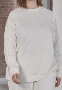 Felpa bianca a maniche lunghe con costine e vestibilità ampia. Presenta sottili righe verticali e strisce di accento sulle maniche. Texture morbida, design semplice.