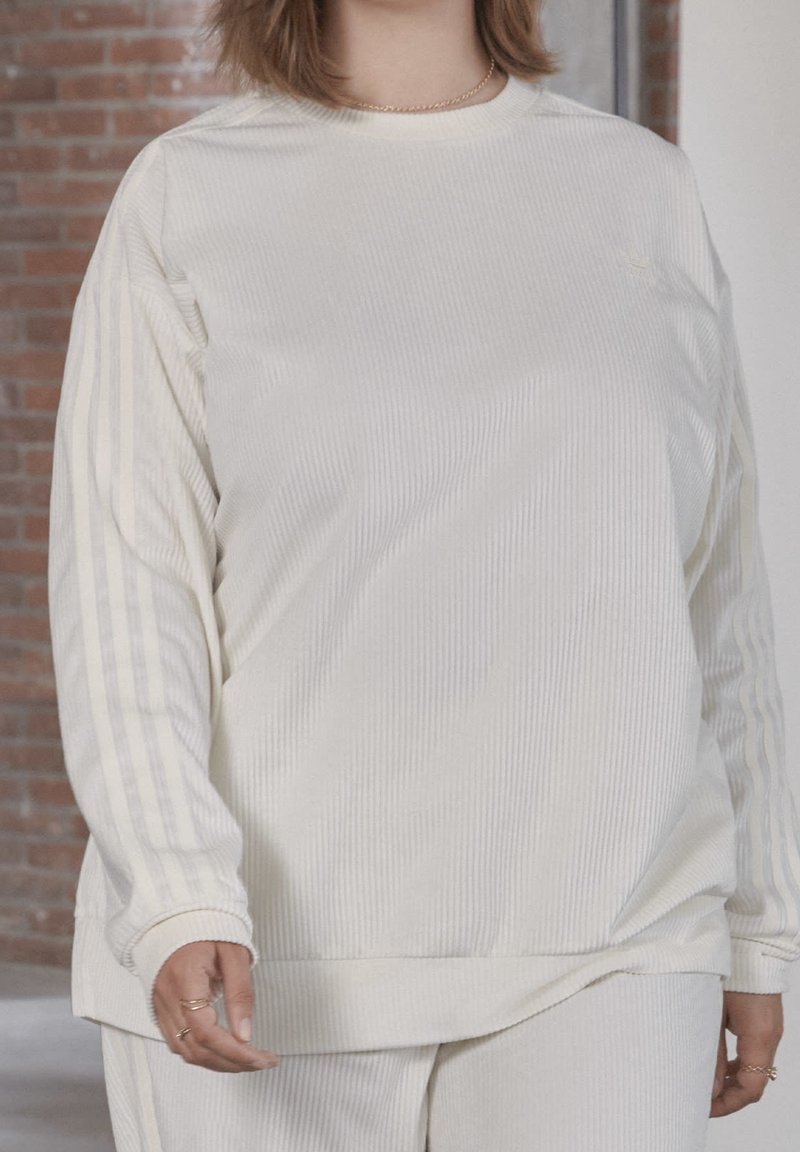 Felpa bianca a maniche lunghe con costine e vestibilità ampia. Presenta sottili righe verticali e strisce di accento sulle maniche. Texture morbida, design semplice.