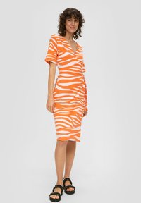 Kleid in Orange und Weiß mit Zebramuster, kurzen Ärmeln, Wickeldesign und seitlichem Bindeband. Schwarze Plateausandalen mit geflochtenen Riemen.