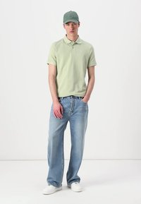 Pólo verde claro, jeans azuis desbotados e ténis brancos; modelo a usar um boné verde a condizer, de pé com as mãos nos bolsos.