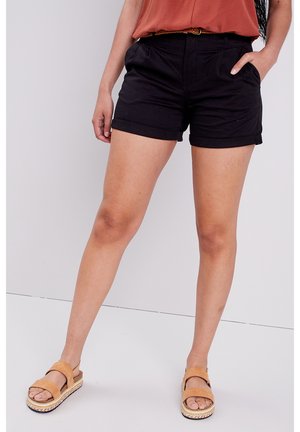 Black tailored shorts with rolled cuffs, featuring side pockets and a smooth texture. Paired with tan sandals with a braided sole.