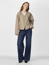 Jachetă bej, tip wrap, dintr-un material texturat, cu închidere printr-un singur buton, asortată cu blugi dark blue, largi, și pantofi cu toc ascuțit negri.