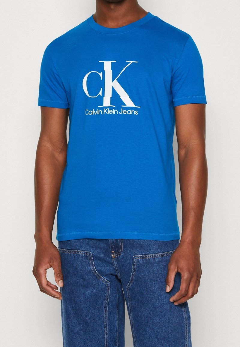 Modrá bavlněná tričko s bílým logem "CK" a textem "Calvin Klein Jeans". Klasický kulatý výstřih a krátké rukávy. Měkká textura.