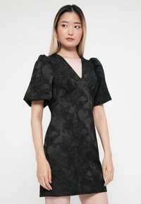 Čierne šaty s kvetinovou textúrou, hlbokým výstrihom v tvare V, balónovými rukávmi, priliehavým strihom a dĺžkou nad kolená. Žiadne viditeľné hardvérové prvky ani akcenty.