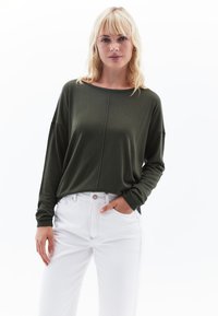 Olijfgroene top met lange mouwen, brede halslijn, ontspannen pasvorm en omgeslagen mouwen, gecombineerd met witte jeans met voorzakken.