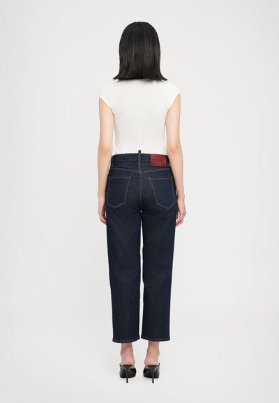 Jeans din denim albastru închis cu croială dreaptă, cu o etichetă pe spate și două buzunare în spate; purtate cu un top alb ajustat și pantofi cu toc negri.