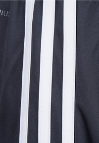 Donkere stof met drie verticale witte strepen en stikdetails, die een sportief ontwerp benadrukken. Logo aanwezig aan de bovenlinkerkant.