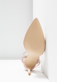 Chaussure à talon haut rose pâle avec un bout pointu, semelle texturée et brides ornées de strass. Matériau lisse, design de talon effilé.