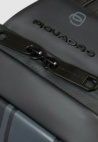 Sac en cuir noir avec une finition brillante, fermeture à double zip, logo embossé et accent contrastant à rayures grises sur le côté.