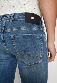 Primer plano de una persona que lleva puestos unos jeans de mezclilla azules con una etiqueta negra de la marca LTB en la cintura y una camisa blanca con estampado metida por dentro.