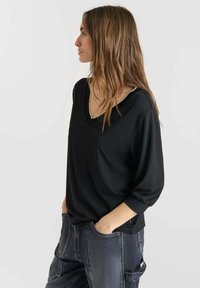 Donna con lunghi capelli castani che indossa un top nero a V e jeans grigio scuro, con le mani nelle tasche, rivolta verso sinistra su uno sfondo neutro.
