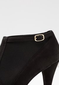 Svart högklackad sko med överdel i mocka och elastisk. Sko har en smal rem och detaljer med metallspänne vid vristen.