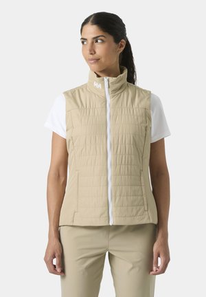 Kobieta ubrana w beżową pikowaną kamizelkę bez rękawów na białą koszulkę z krótkim rękawem, patrząca na bok, z rękami swobodnie opuszczonymi wzdłuż ciała.