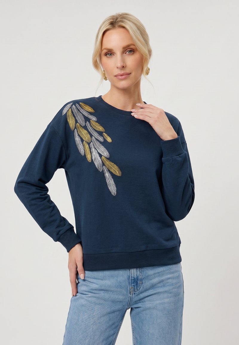 Femme blonde portant un sweat-shirt bleu marine avec une broderie de feuilles dorées et argentées sur l'épaule et un jean bleu clair.