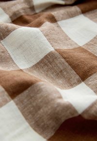Tissu écossais marron et blanc avec une texture tissée, présenté avec des plis doux et des ombres légères.