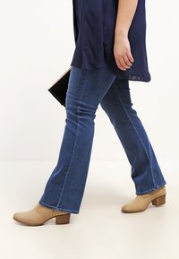 Botines de tacón beige con un upper texturizado, combinados con jeans acampanados de color azul oscuro. La parte superior es una camisa suelta de color navy que cubre parcialmente los jeans.