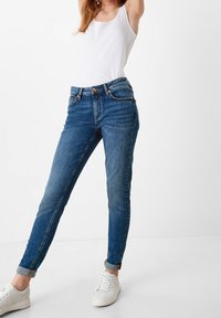 Blue denim jeans with a slim fit, featuring a mid-rise waist, five pockets, and rolled cuffs. Worn with a white tank top and white sneakers.