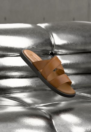 Brown leather slide sandal with two wide straps and black sole floating above shiny metallic silver tufted surface.