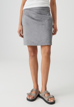 Frau trägt einen knielangen grauen Wildlederrock, ein weißes ärmelloses Oberteil und graue Sandalen mit doppelter Schnalle, steht auf einem schlichten hellen Hintergrund.